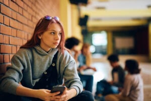 Pensive teenage girl using cell phone at high school hallway and looking away.