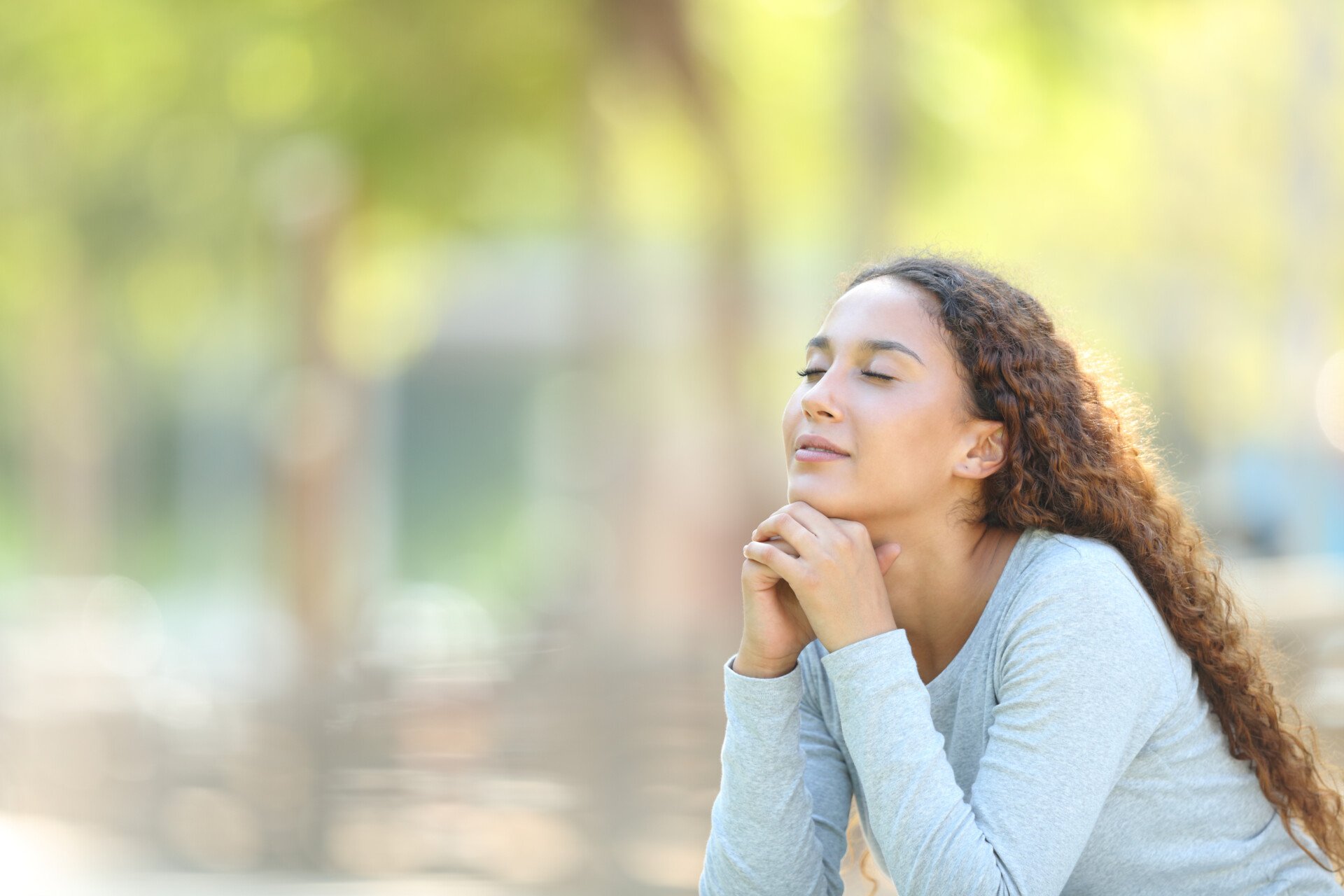 a woman out in nature with her eyes closed, resting chin on hands, demonstrating mindfulness