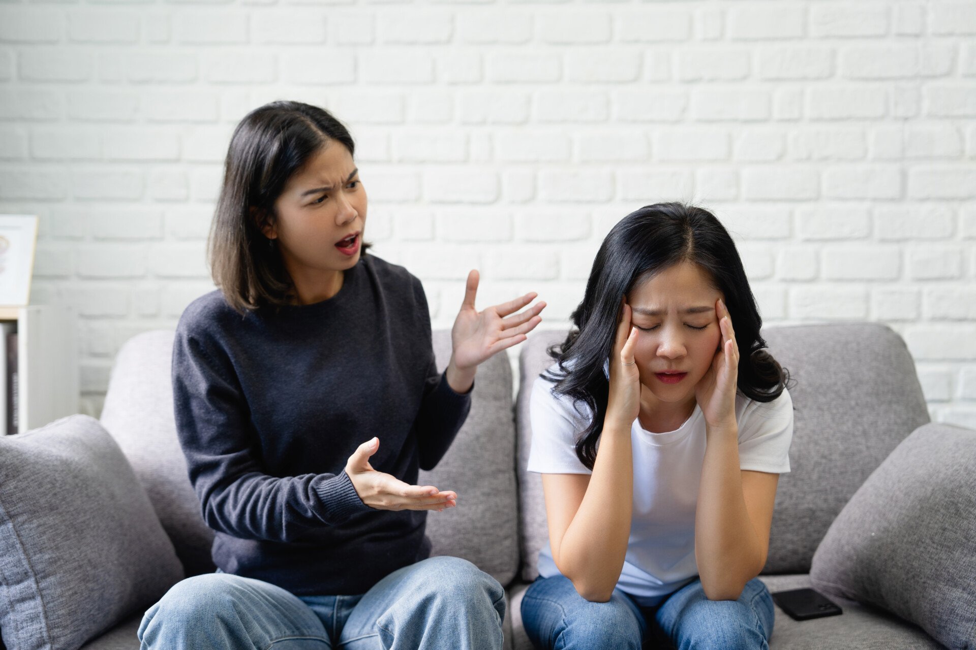 two women arguing on a sofa, one is shouting while the other has head in her hands looking frustrated.